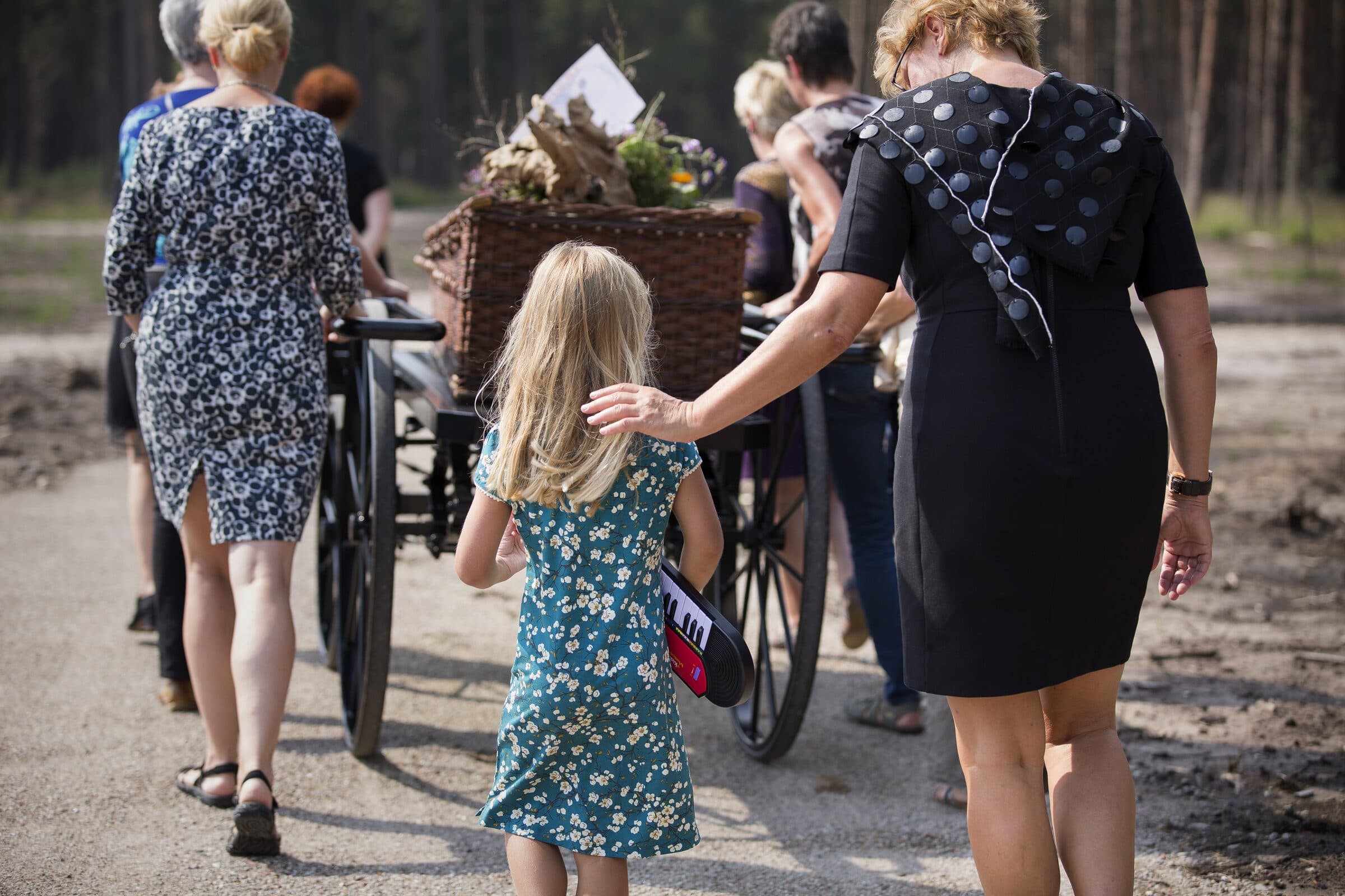 Familie en kinderen lopen samen achter de loopkoets met een rieten kist, een meisje loopt naast haar moeder